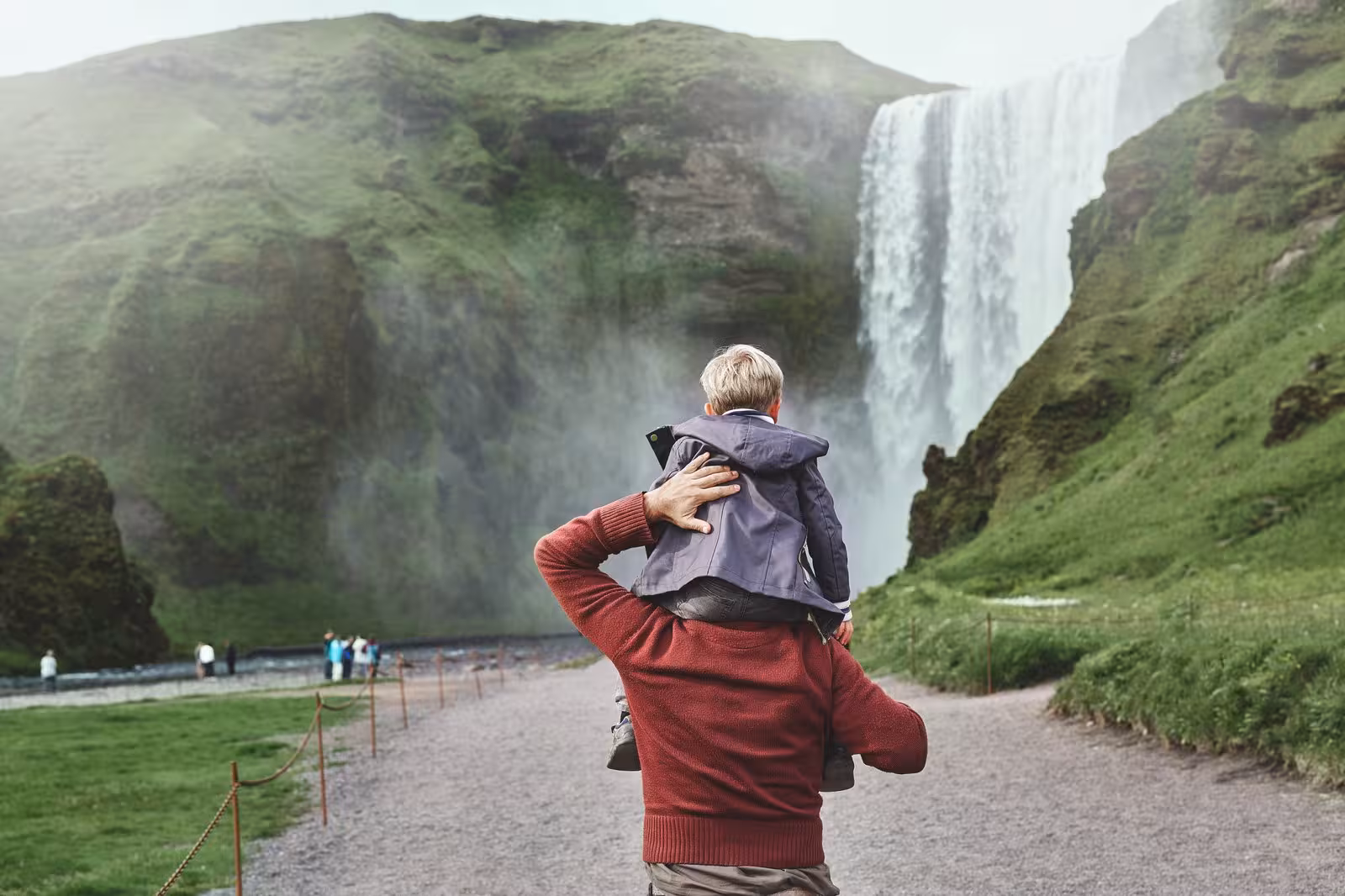 Father and child admire the majestic Skógafoss waterfall on a green Icelandic landscape during a self-drive tour.