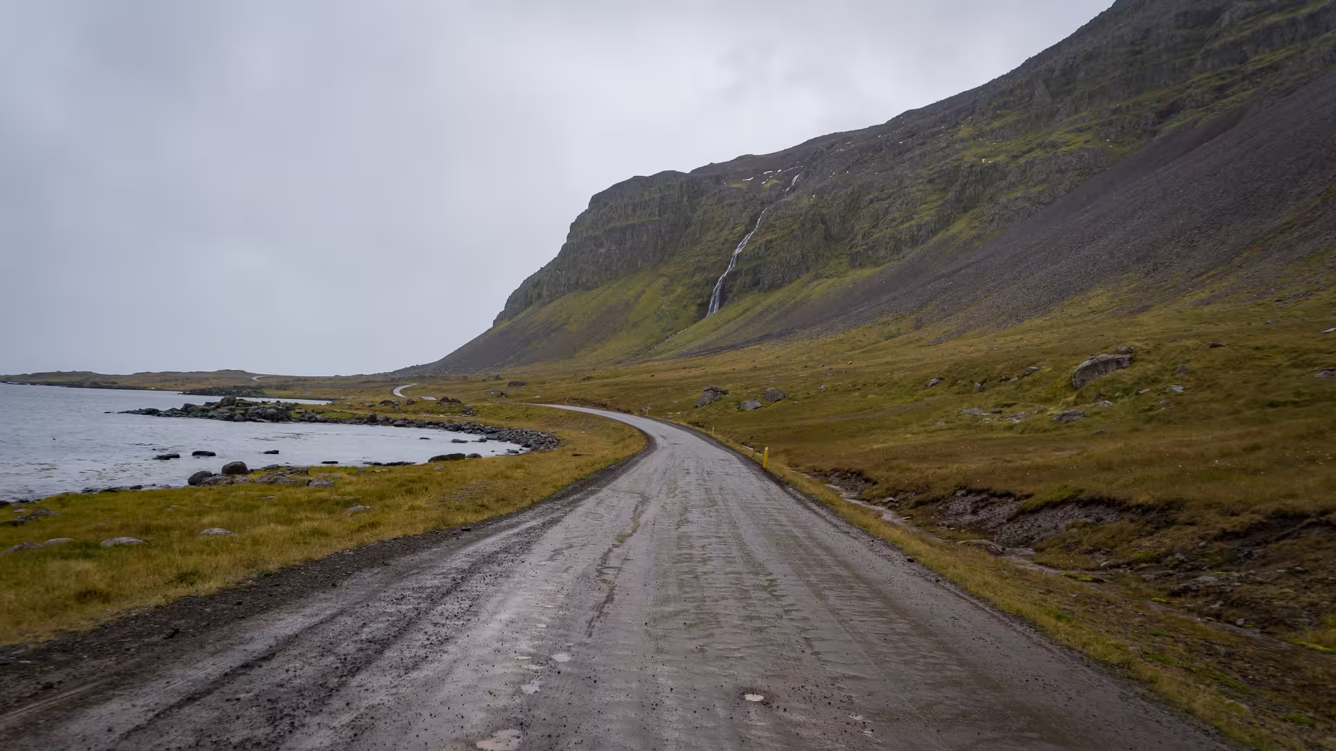 Scenic winding gravel road along Iceland's mystical coastline with dramatic green cliffs and waterfall