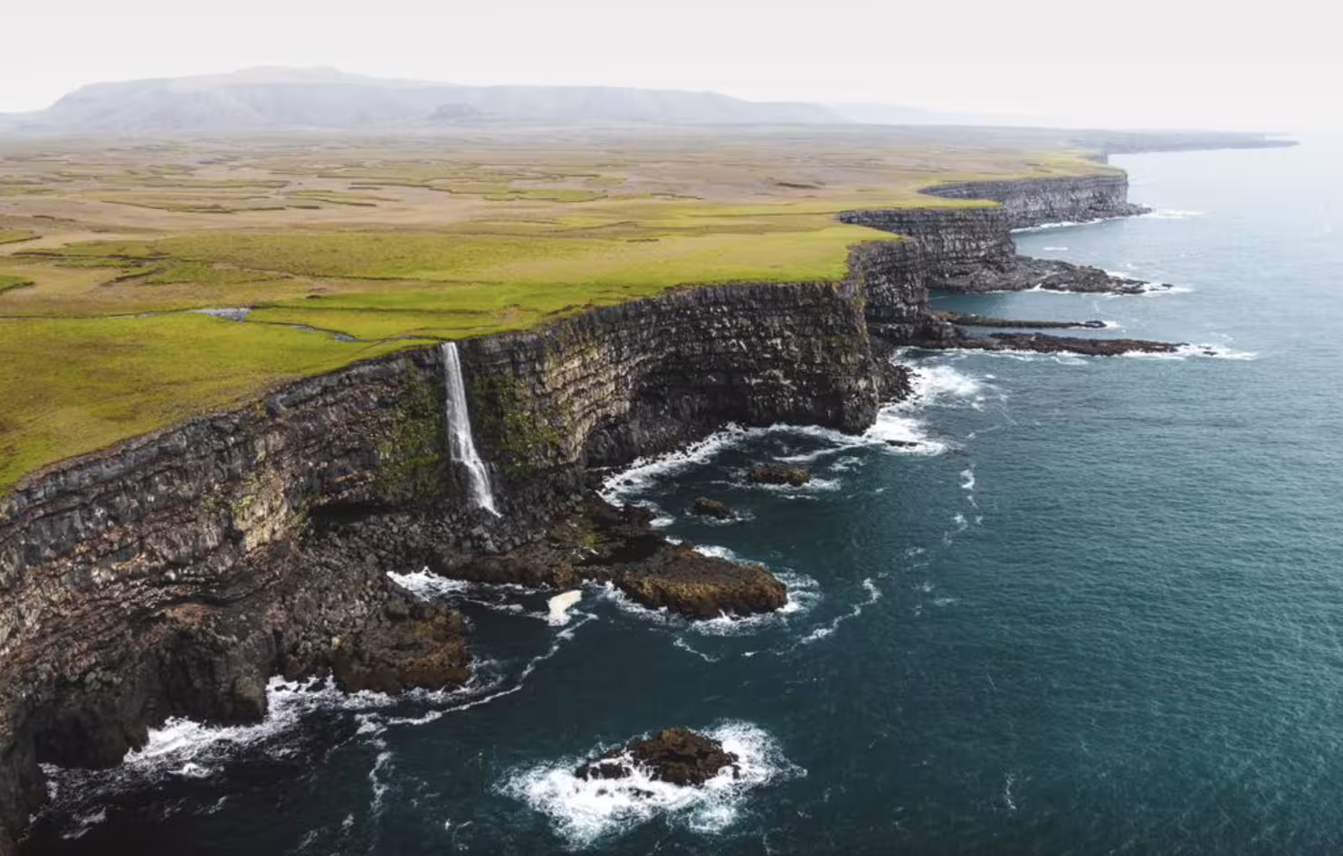Aerial view of dramatic basalt sea cliffs with waterfall dropping into ocean, green Icelandic landscape above