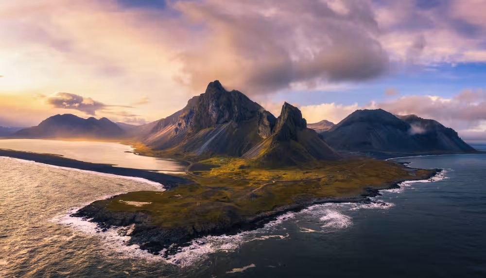 Aerial view of dramatic Icelandic coastal mountains with green slopes, black sand beaches, and golden sunset sky
