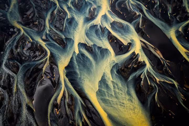 Aerial drone view of braided glacial river channels with teal and yellow hues on black volcanic sand in Iceland
