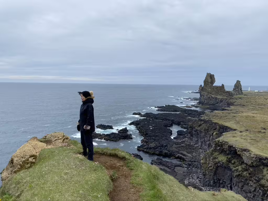Person in winter clothing standing on coastal cliffs overlooking dramatic Iceland sea stacks and ocean