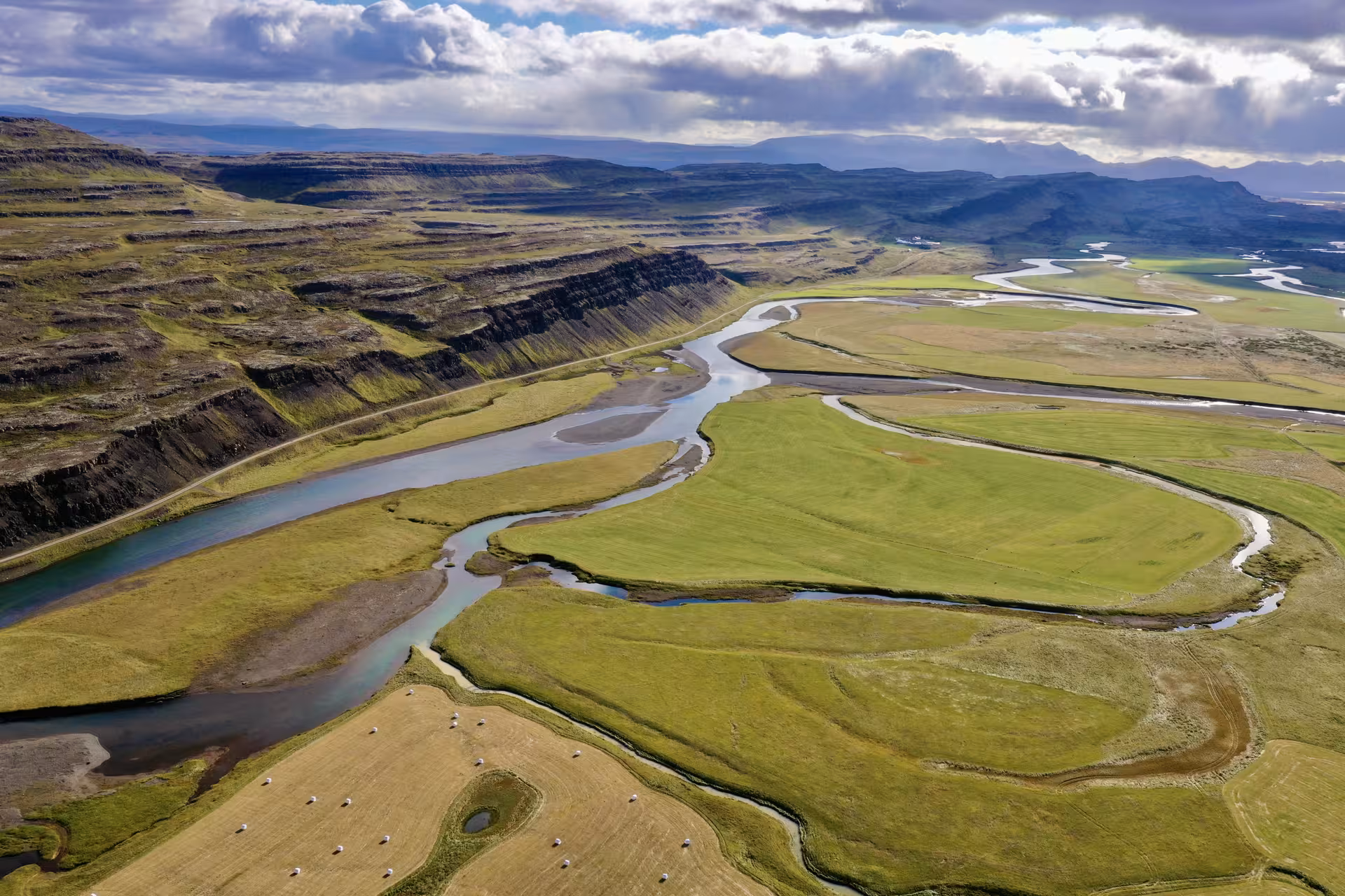 Aerial view of a winding glacial river through green Icelandic farmland with layered basalt cliffs
