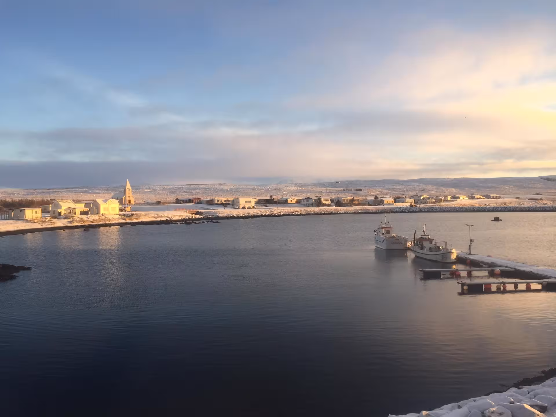 Snowy Icelandic harbor town at golden hour with fishing boats docked at pier and snow-covered hills