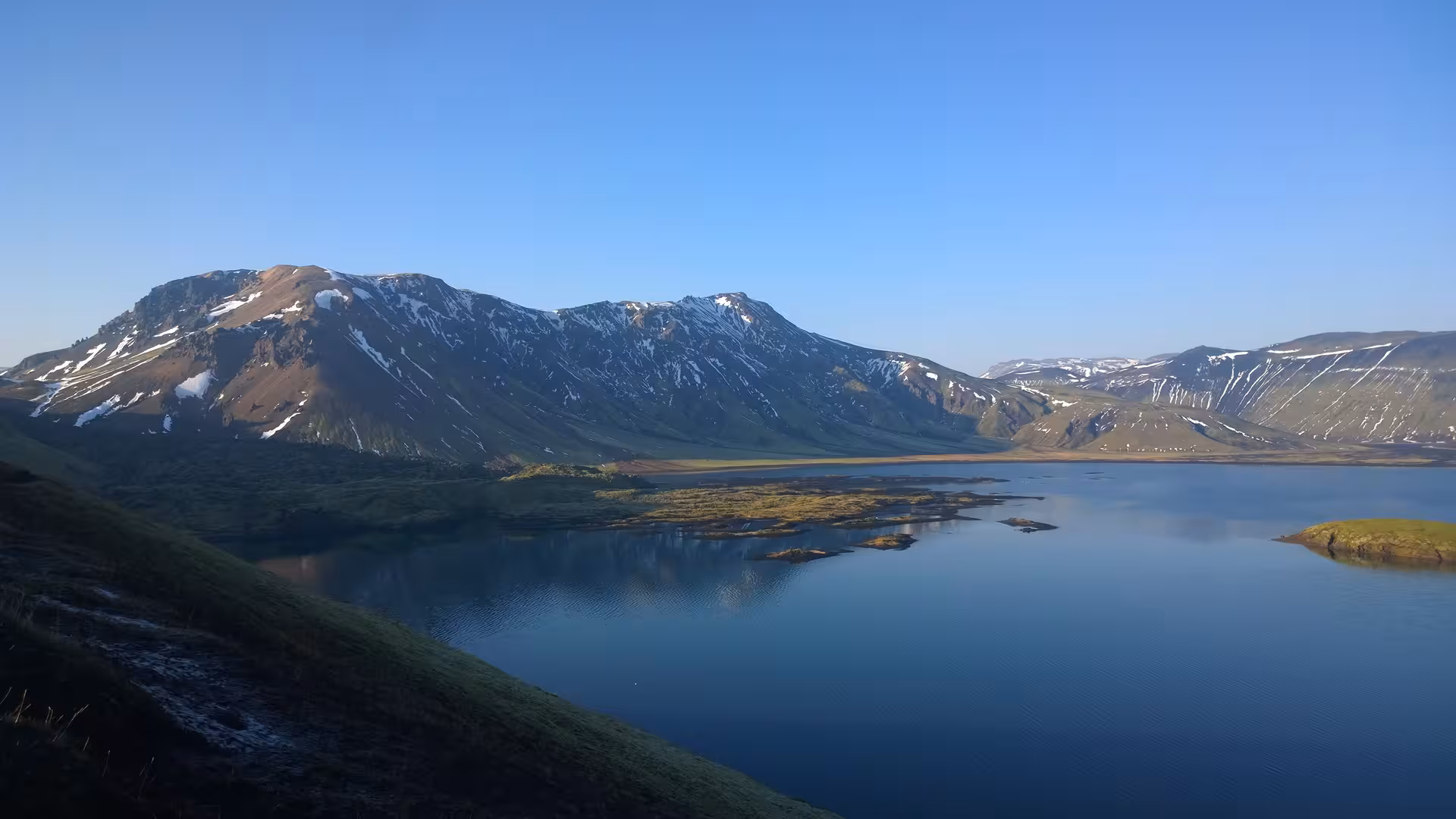 Calm glacial lake reflecting snow-capped mountains under clear blue sky in Iceland's Fjallabak highlands