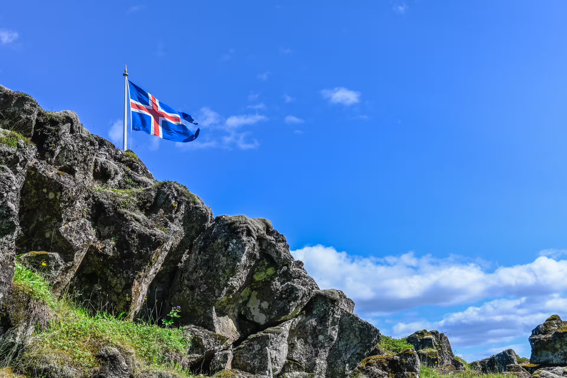 Icelandic flag waving atop rugged lava rock formation under a bright blue sky in Iceland