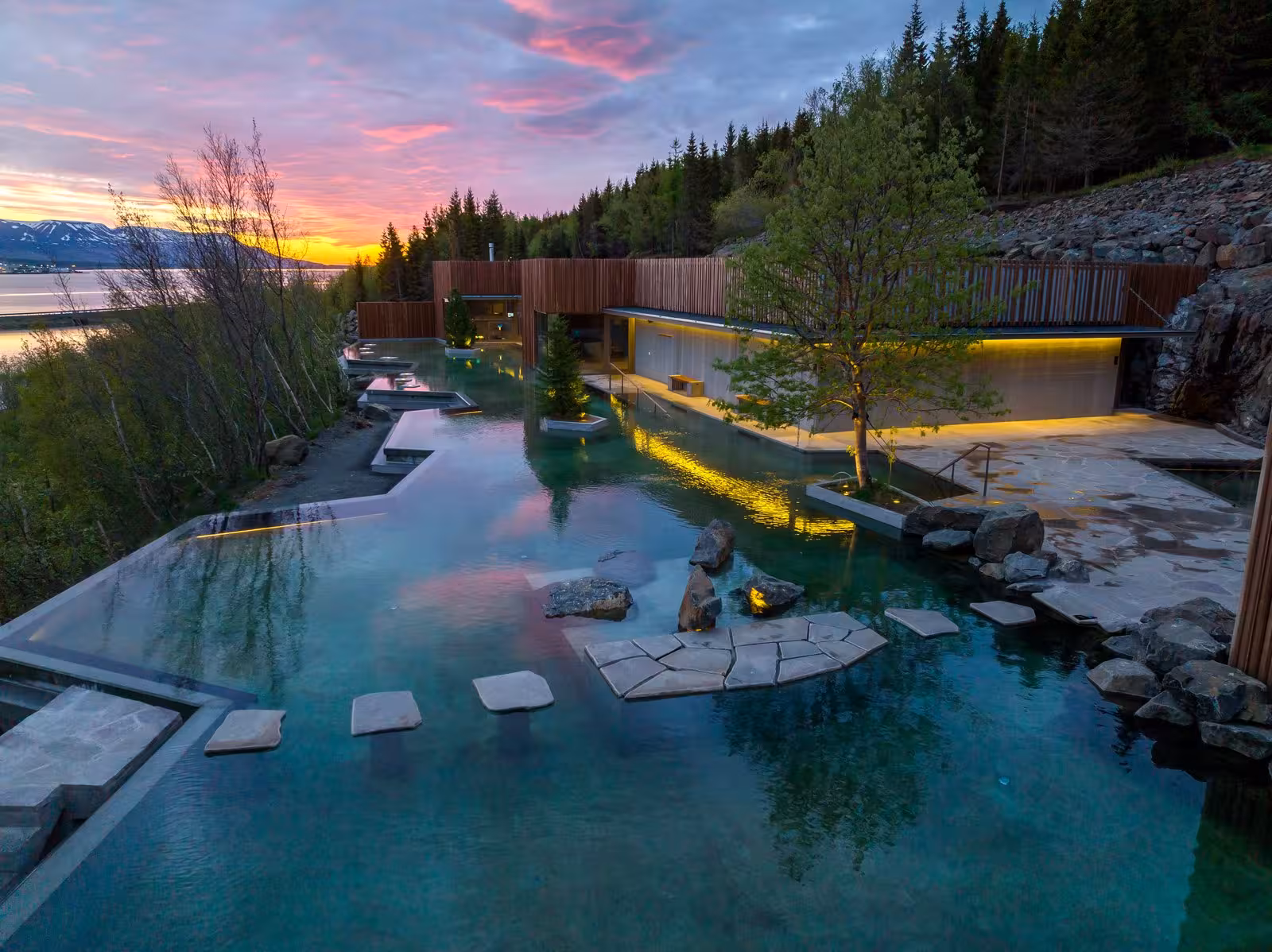 Sunset over a serene Icelandic geothermal spa surrounded by lush forest and calm waters.