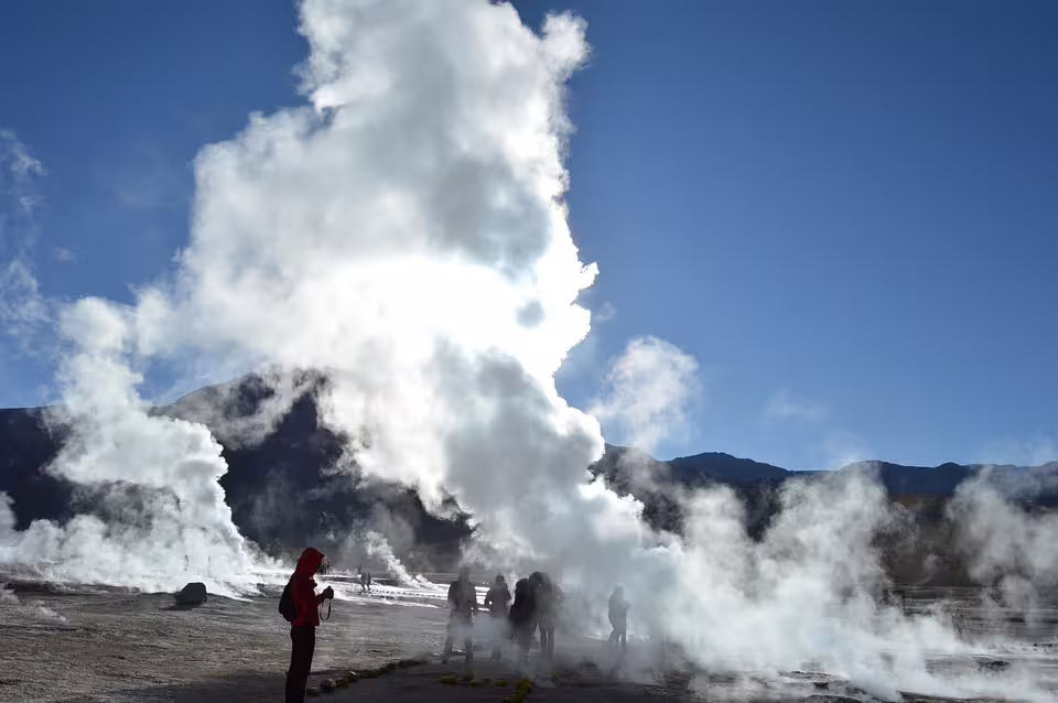 Tourists watching massive geothermal steam vents erupt against a clear blue sky with mountains in background
