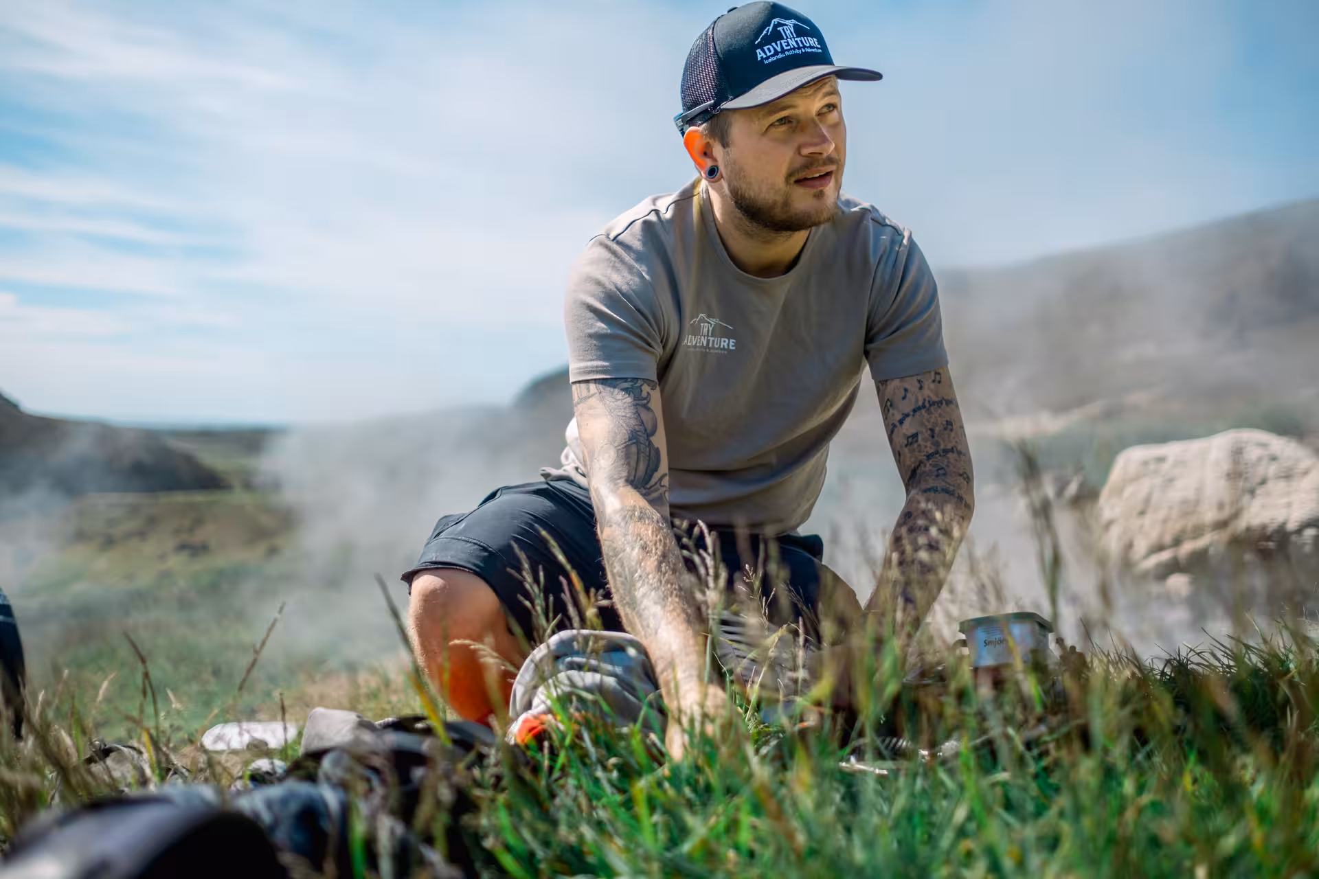Tattooed guide kneeling on grass in Iceland geothermal valley with steam vents near Hengill Volcano