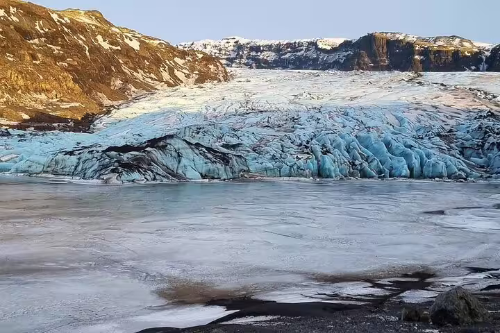 Blue glacier ice face with crevasses meeting a frozen lagoon, surrounded by volcanic rock mountains in Iceland