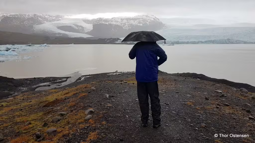 Traveler with umbrella overlooking Jökulsárlón glacier lagoon with icebergs on a rainy day in Iceland