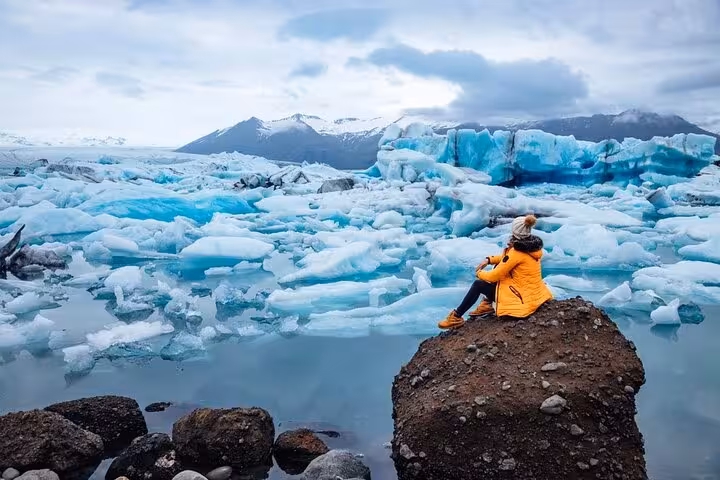 Traveler in yellow jacket sitting on rock overlooking Jökulsárlón Glacier Lagoon with blue icebergs, Iceland