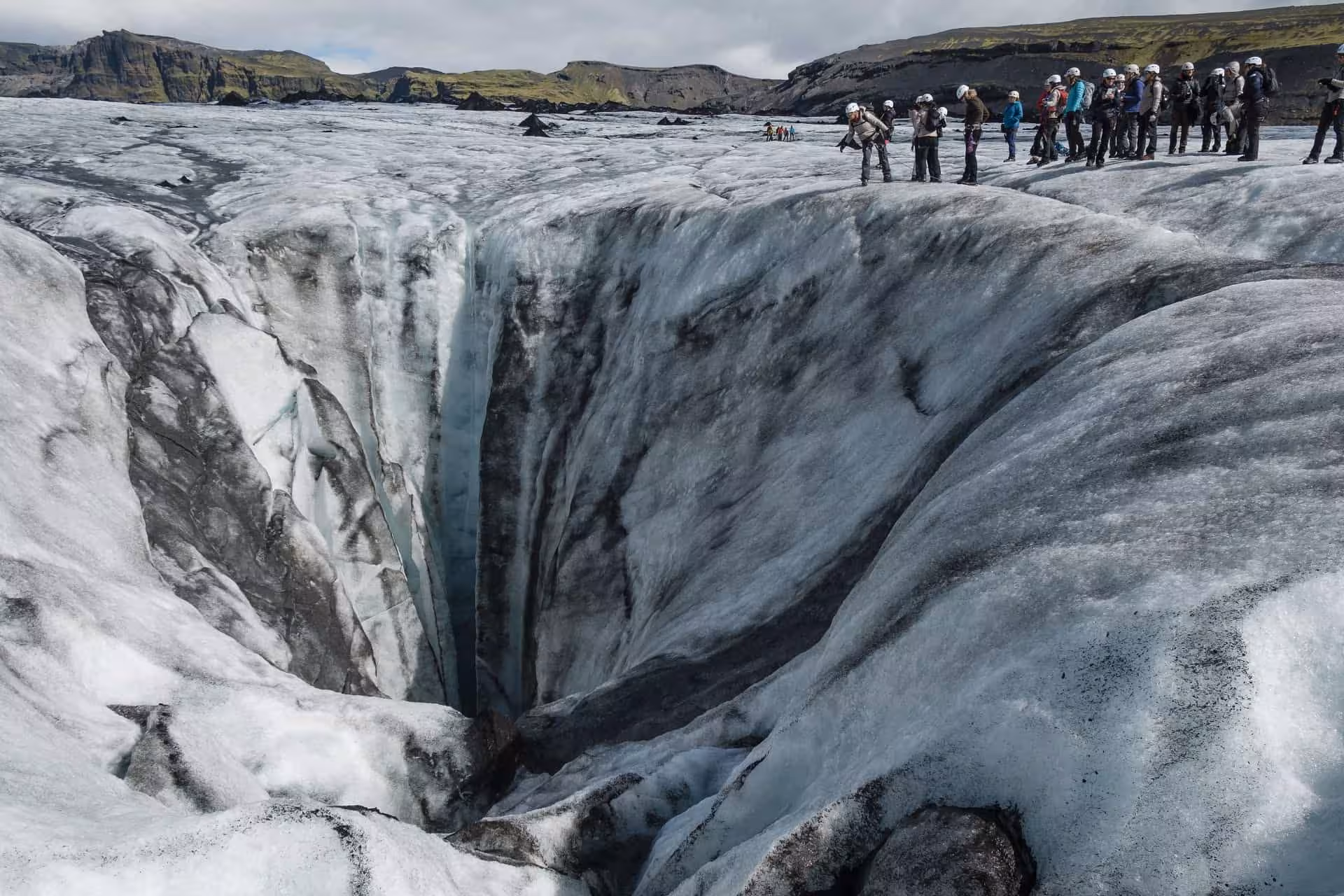 Tour group in helmets standing at edge of deep glacial crevasse on Iceland's Sólheimajökull glacier