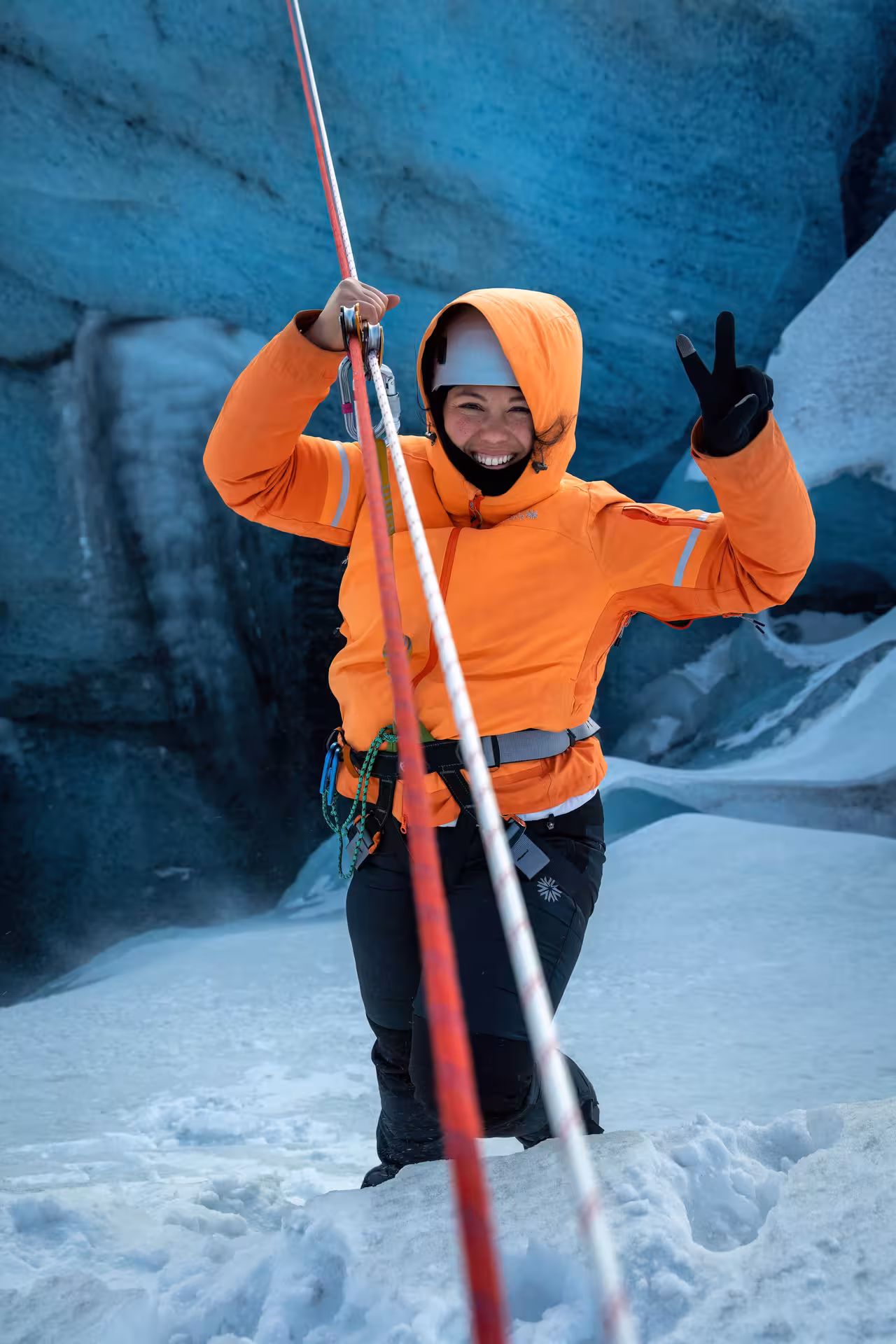 A happy adventurer in full safety harness and orange jacket poses on an Iceland glacier zip line guided winter tour.