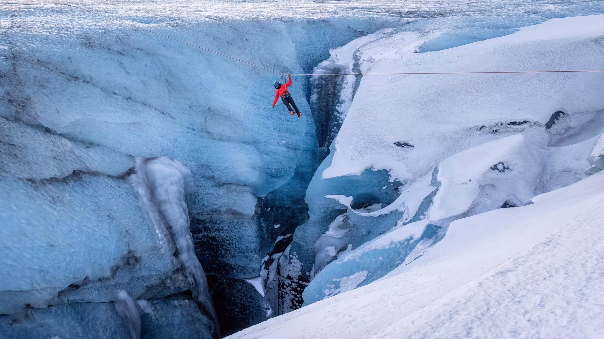 A thrilling glacier zip line adventure in Iceland with a participant in a red jacket soaring over a deep blue ice crevasse in winter.