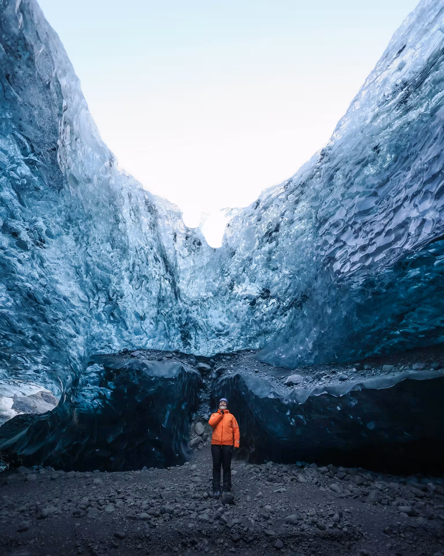 Adventurer in orange jacket gazes up at towering blue glacier ice walls during a guided winter zip line tour in Iceland.