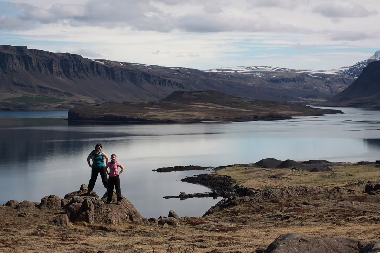Two hikers standing on rocks beside a calm fjord with dramatic cliffs and snow-capped mountains in Iceland