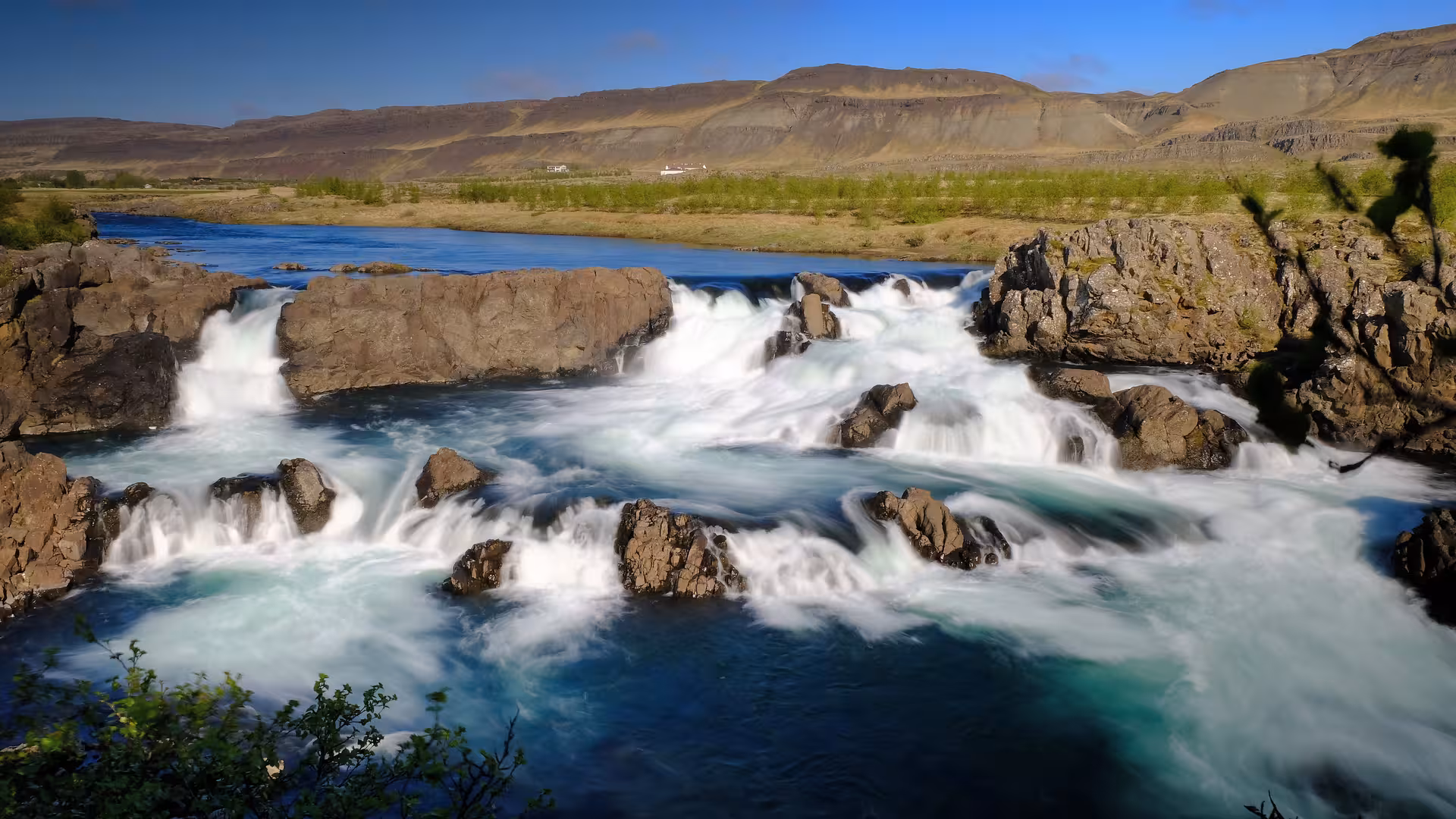 Rushing turquoise glacial river rapids over basalt rocks with Icelandic mountains and blue sky