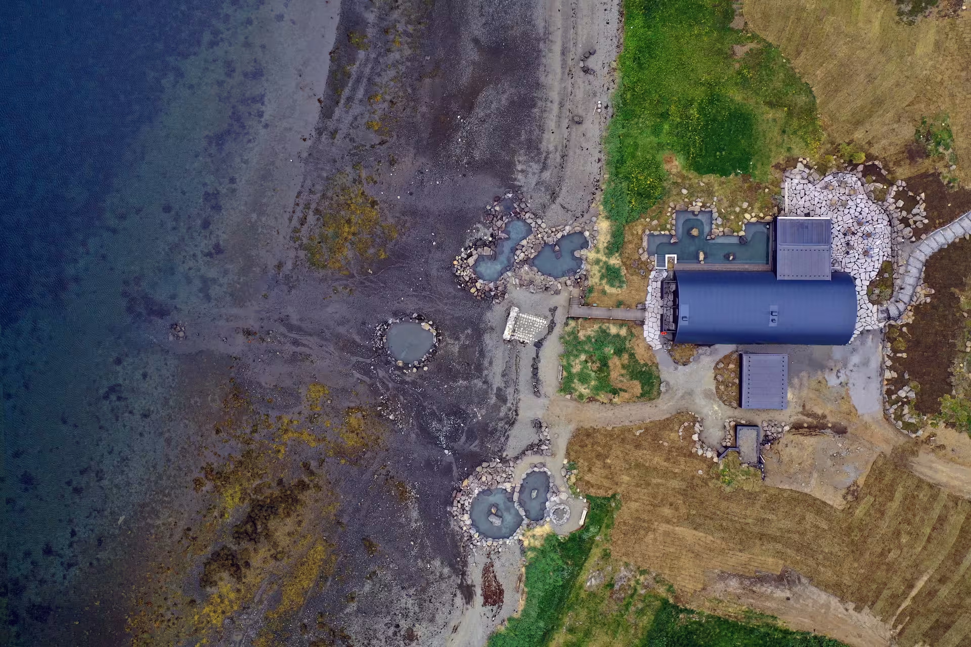 Aerial view of geothermal hot spring pools along Iceland's rocky coastline near a dark-roofed building