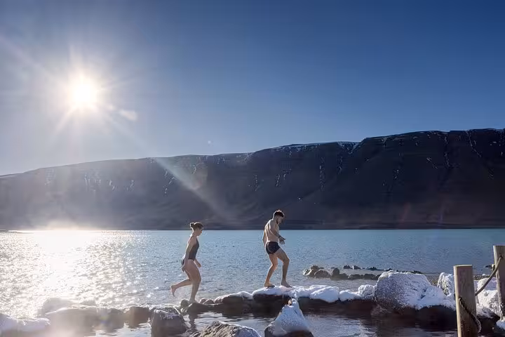 Two people in swimwear stepping on snowy rocks by a glacial lake in Iceland under bright winter sun