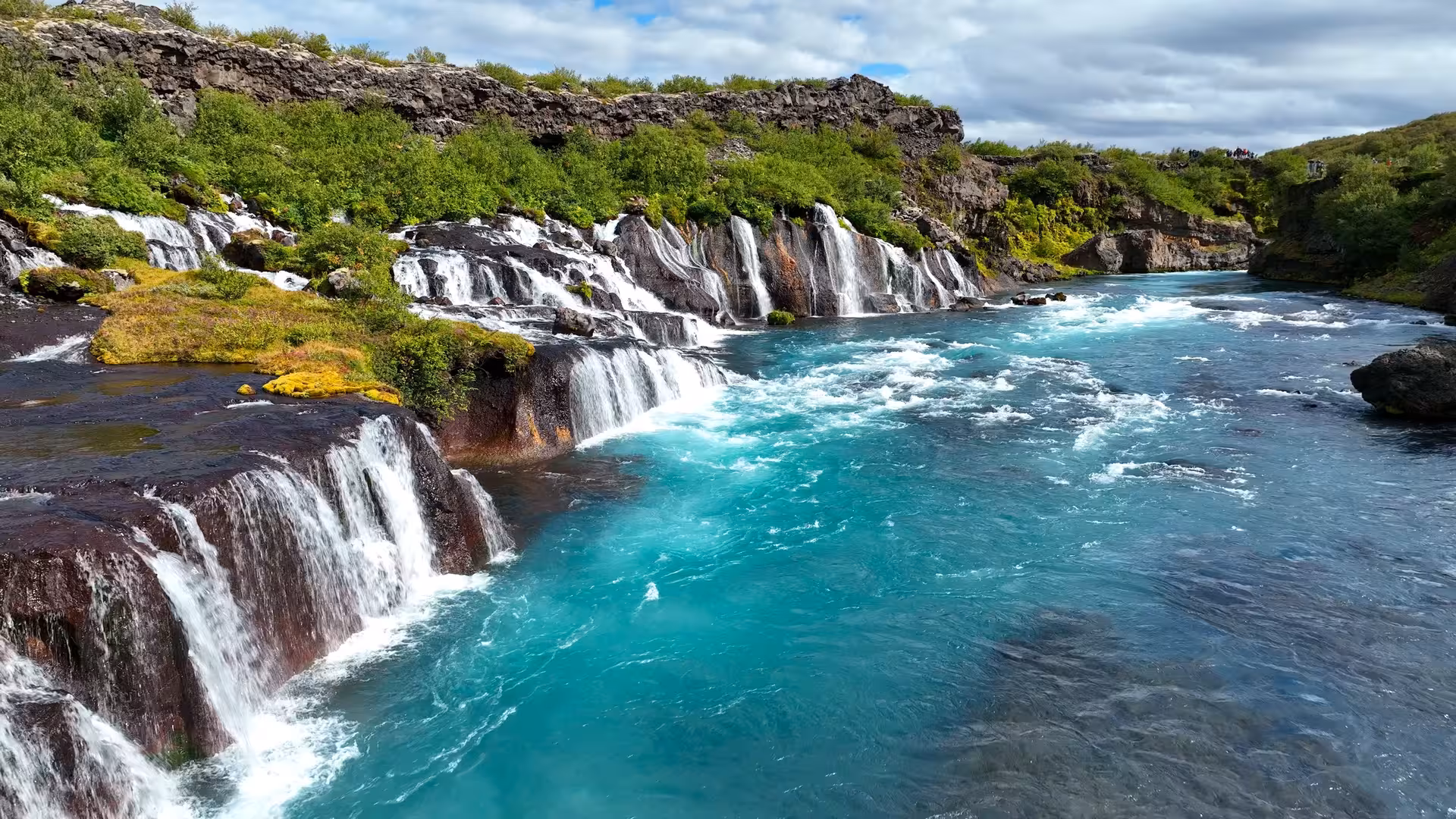 Turquoise glacial river with cascading waterfalls over black lava rocks and green vegetation in Iceland