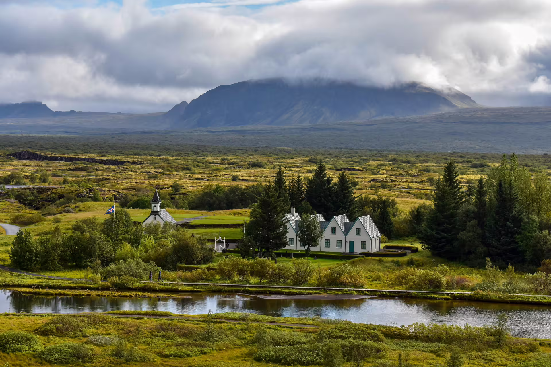 Þingvellir church and houses beside a calm river with volcanic mountains and clouds in Iceland