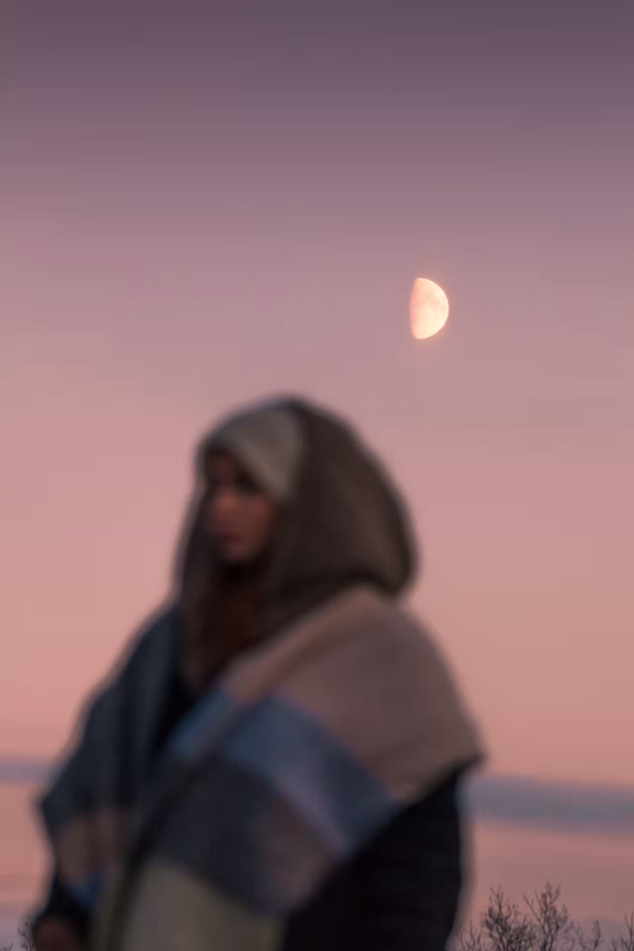 Woman in hooded coat gazing at half moon in pink twilight sky in Iceland
