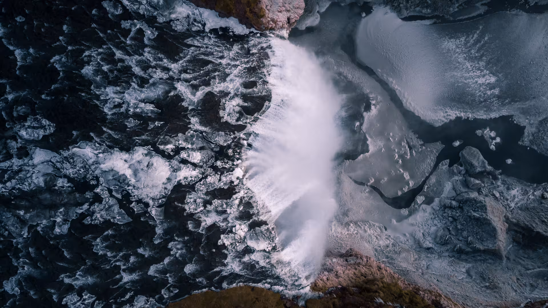 Aerial view of a powerful Icelandic waterfall crashing over dark rocks with surrounding snow and ice