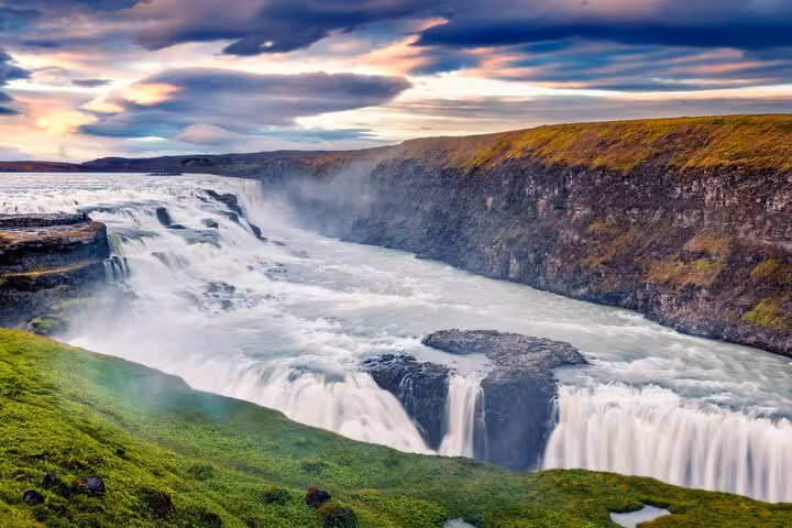 Gullfoss waterfall cascading into a misty canyon with green cliffs and dramatic sunset sky in Iceland