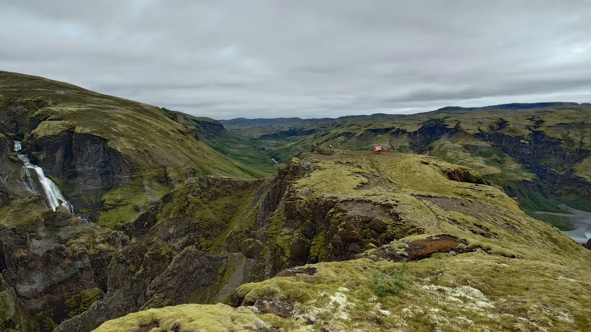 Red helicopter landed on mossy cliffside ridge in Iceland with waterfall and green canyon valley below