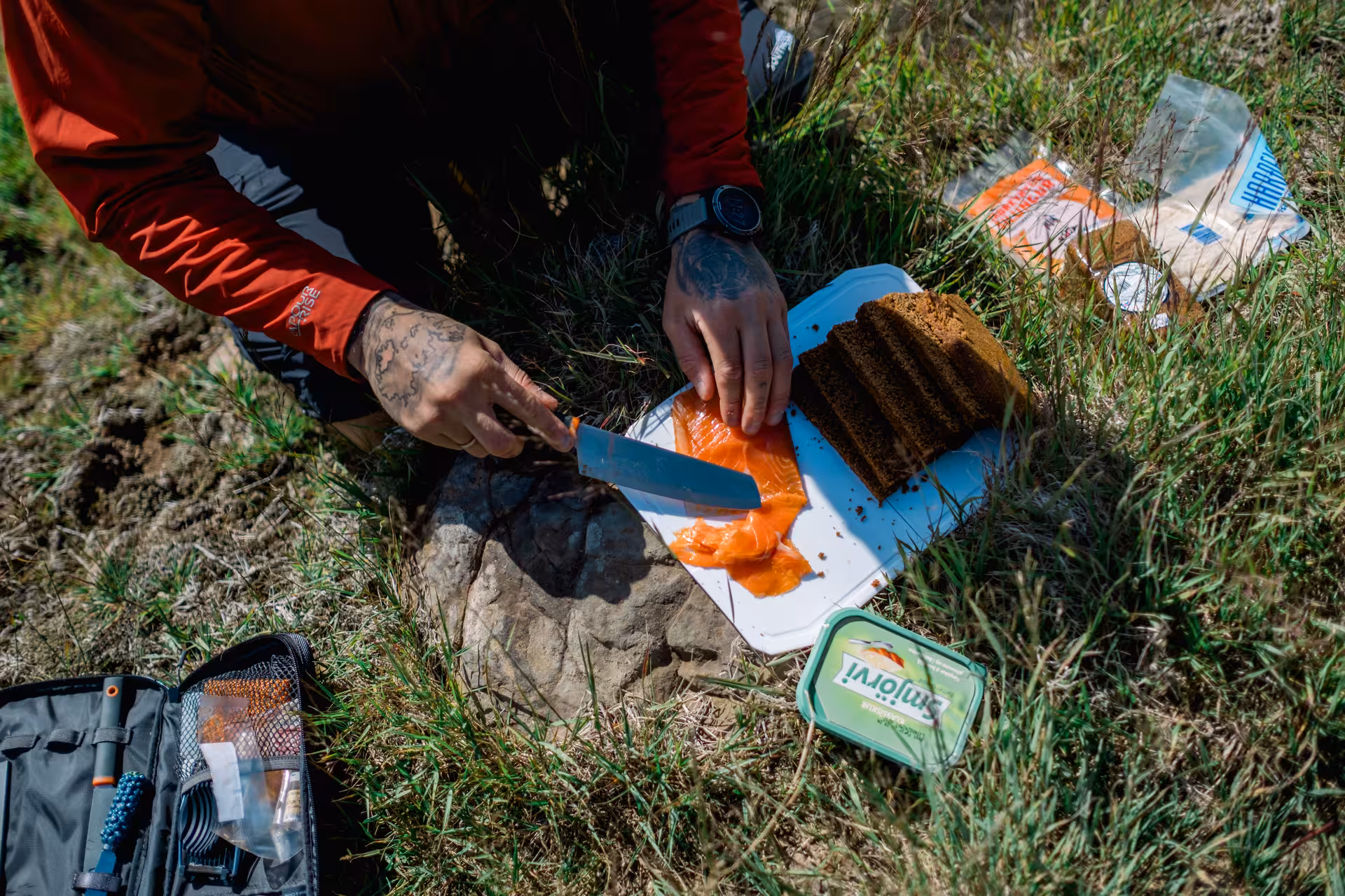 Tattooed hiker slicing smoked salmon with dark rye bread and Smjörvi butter for an outdoor meal in Iceland
