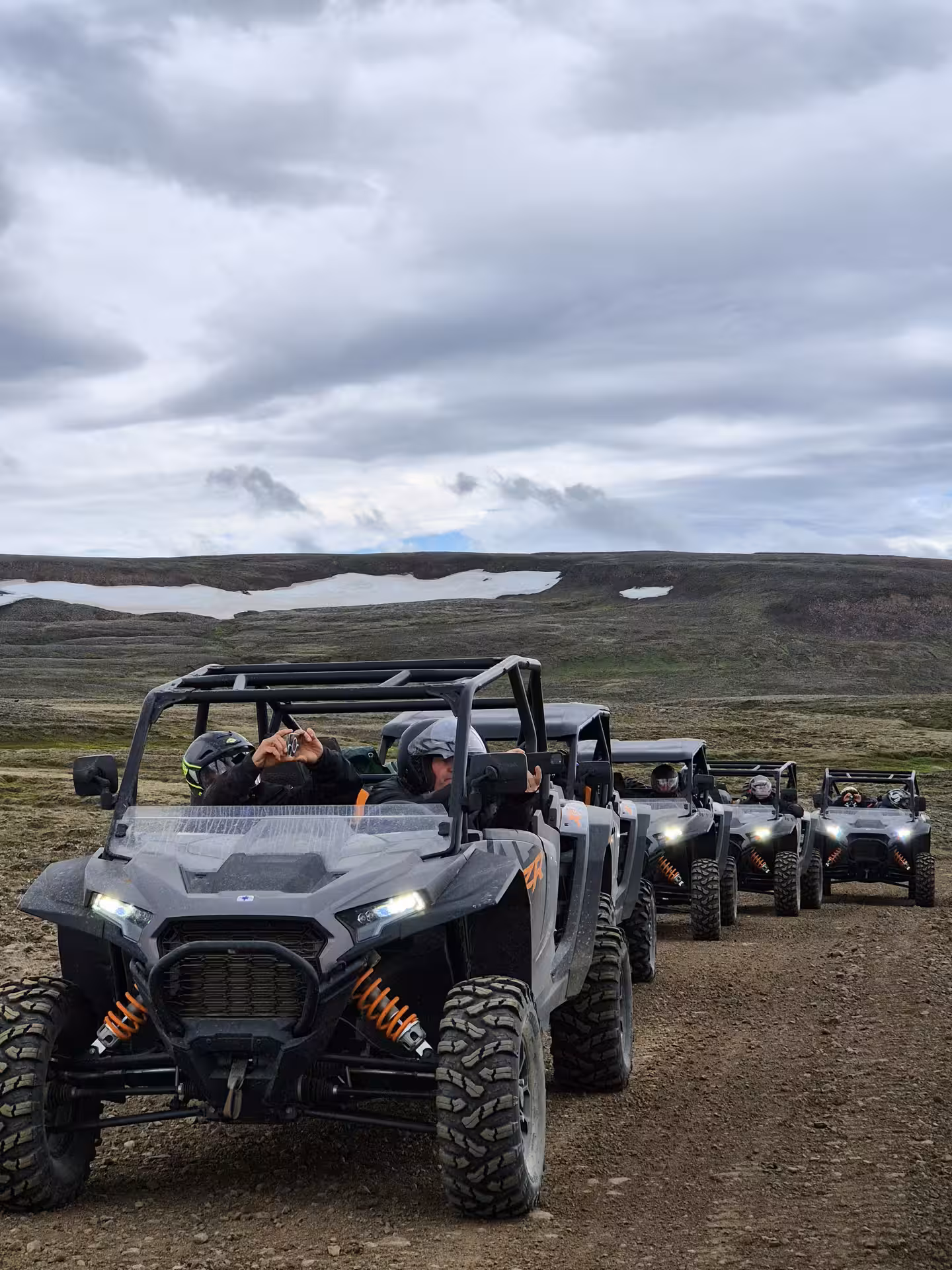 convoy of black and orange buggies on dirt trail through Iceland highlands with snow-capped hills