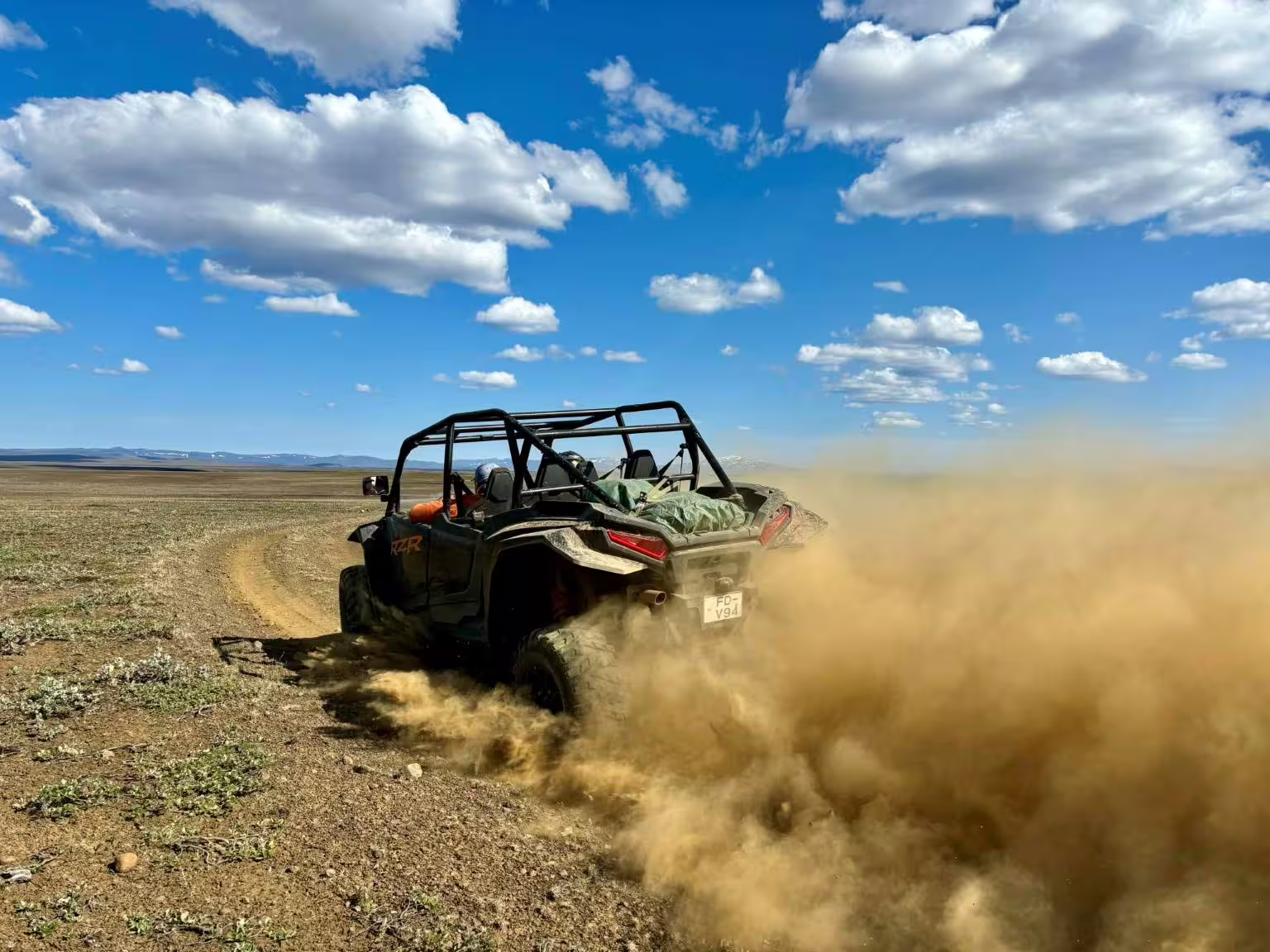 Polaris RZR buggy kicking up dust cloud while driving across open highland terrain under blue sky