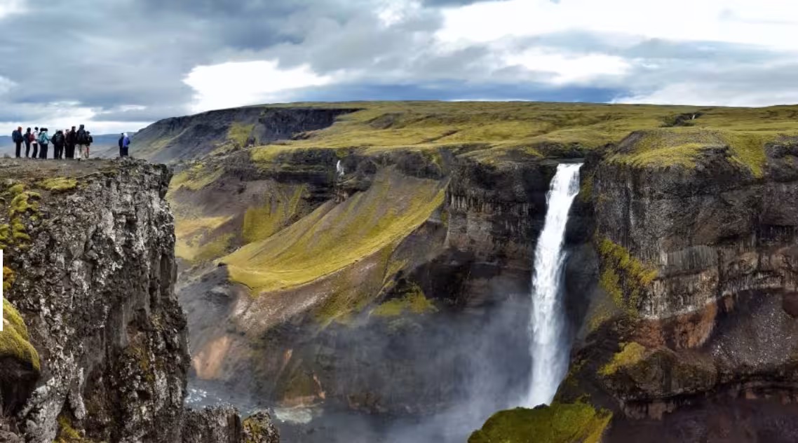 Group of hikers overlooking a dramatic Icelandic highland canyon with a tall waterfall and mossy cliffs