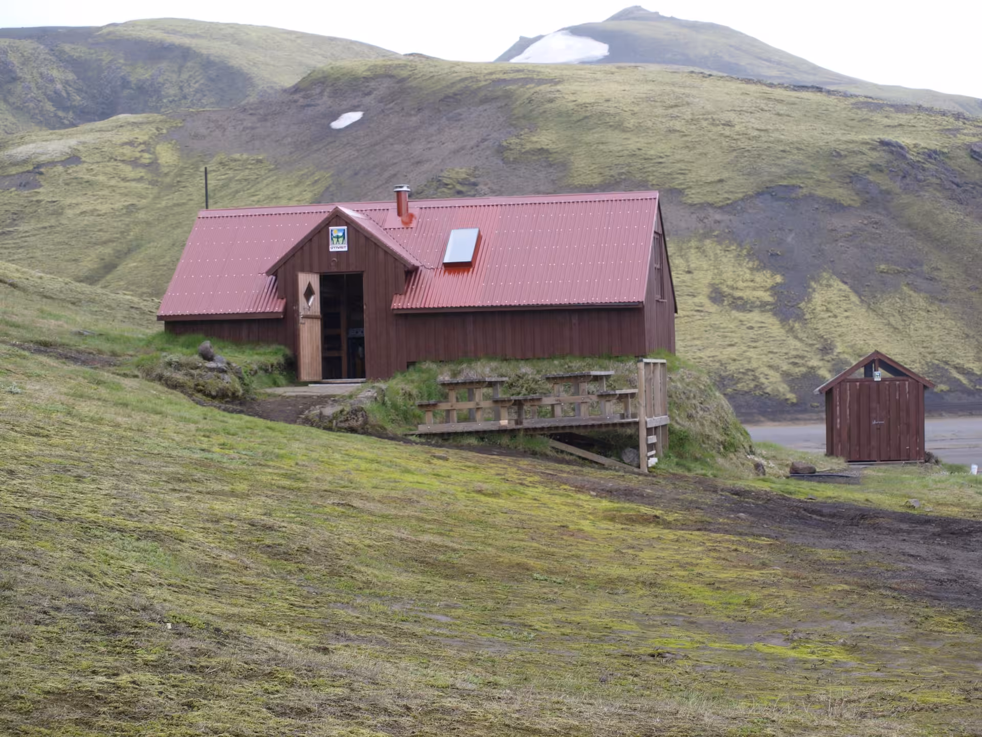 Red-roofed mountain hut on mossy hillside in Iceland Highlands with snow-capped volcanic peaks behind