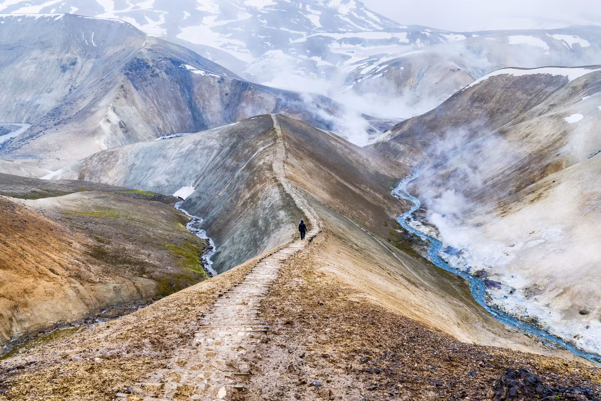 Hiker on stone path ridge in Iceland Highlands with geothermal steam and blue river below