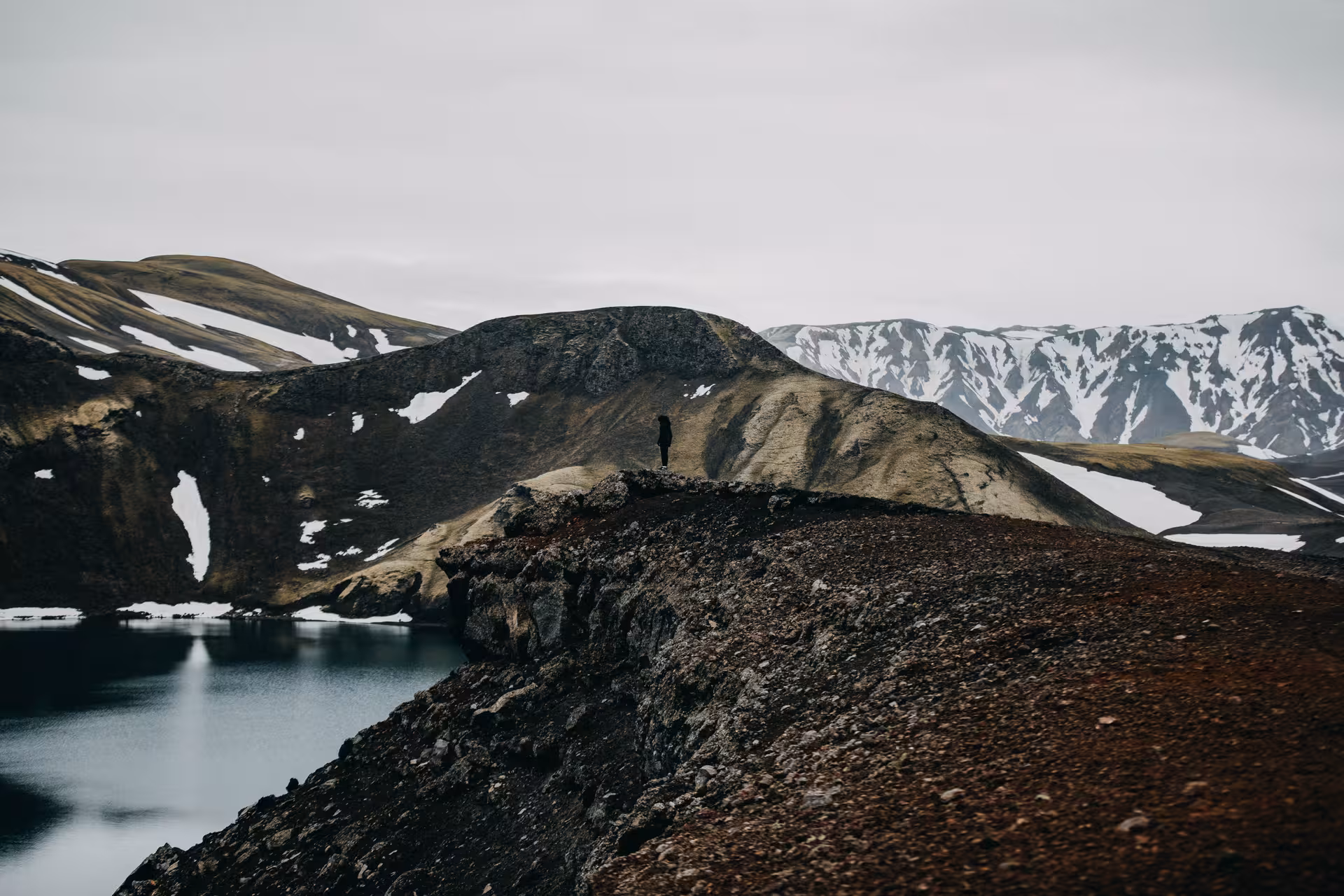 Lone hiker standing on volcanic ridge overlooking glacial lake in Iceland Highlands surrounded by snow-capped mountains