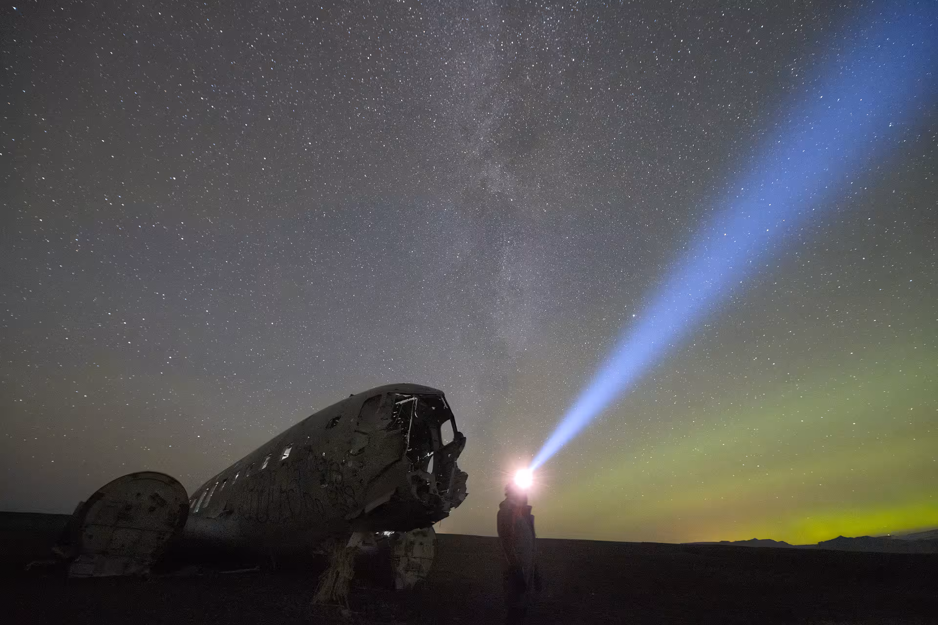 Person with flashlight beam beside wrecked plane under starry sky and northern lights in Iceland highlands