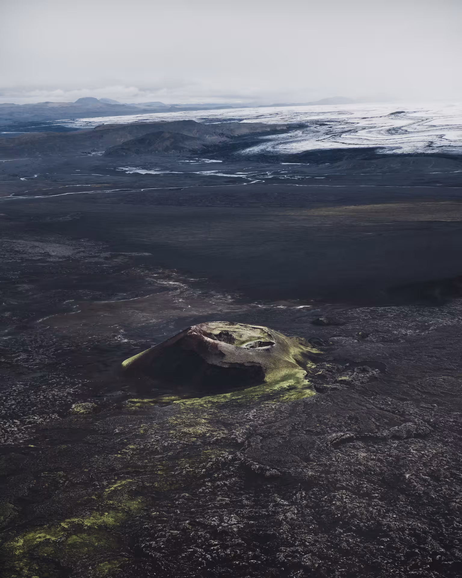 Aerial view of volcanic crater with green moss on black lava fields, glacier in background, Iceland Highlands