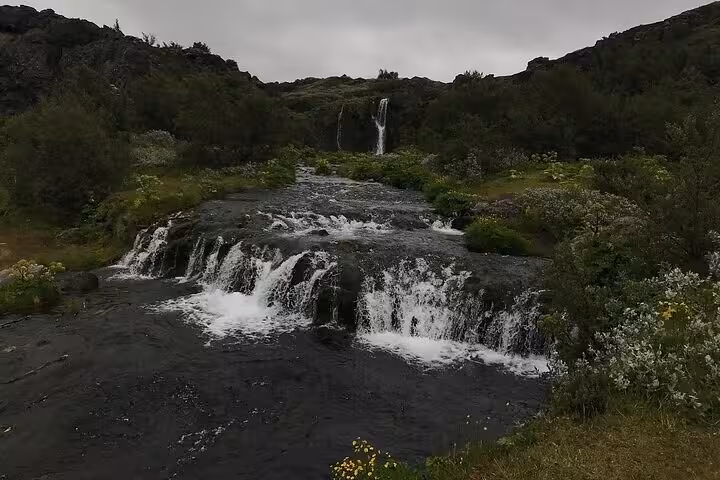 Cascading waterfall over dark volcanic rocks flowing through lush green Iceland highlands landscape