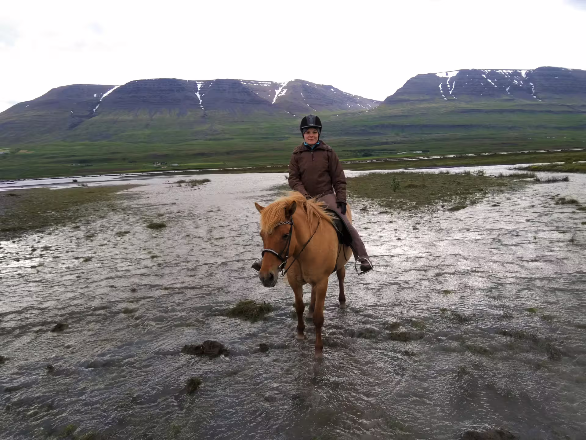 Rider on Icelandic horse crossing shallow glacier river delta with snow-capped mountains in background
