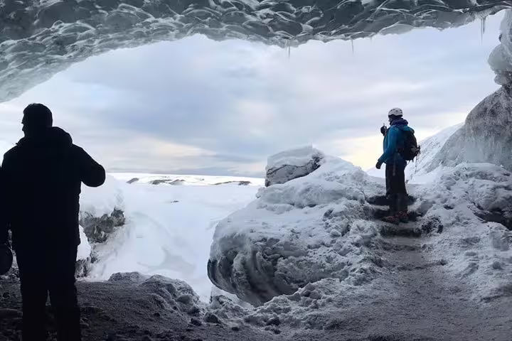 Two hikers exploring an ice cave opening with snowy Arctic landscape and cloudy sky in Iceland