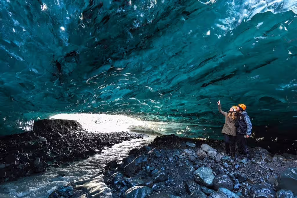 Two tourists in helmets exploring a stunning blue ice cave in Iceland with rocky floor and glacial stream