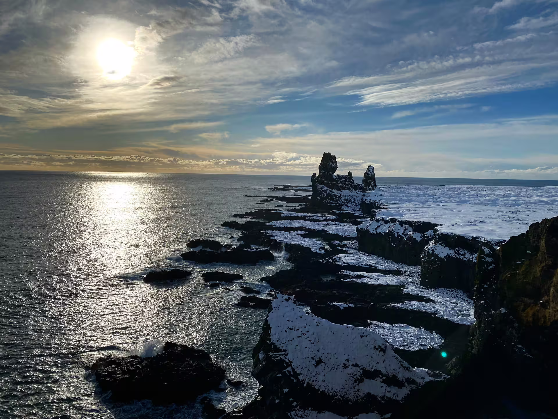 Snow-dusted black lava rock coastline with dramatic sea stacks and sun reflecting on calm ocean waters in Iceland