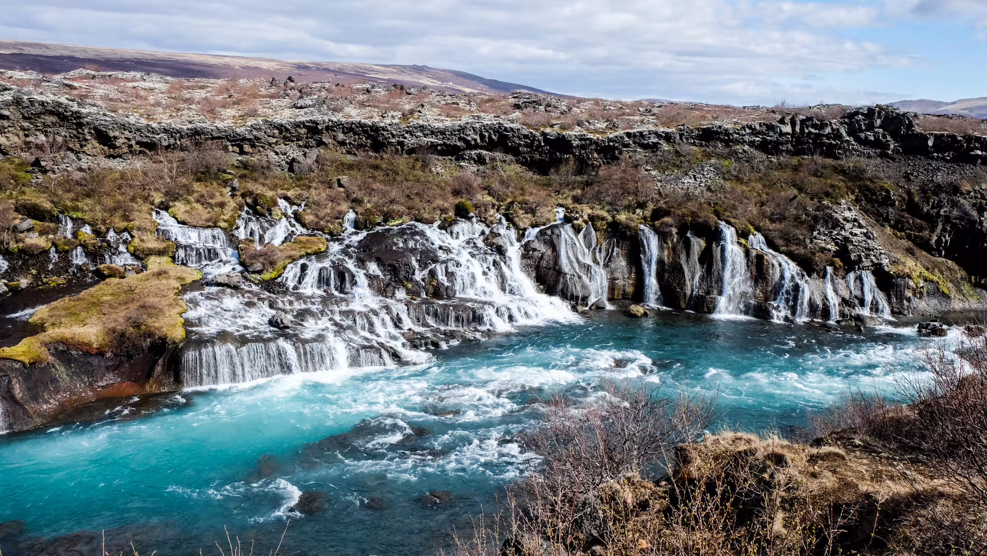 Multiple cascading waterfalls flowing over dark lava rocks into a turquoise river in Iceland