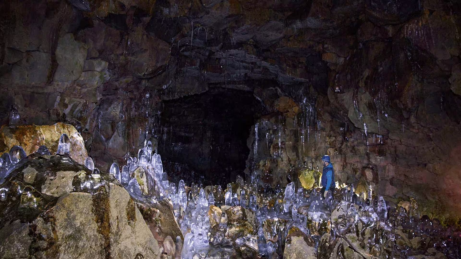 Explorer standing among ice stalagmites inside an illuminated Iceland lava tunnel cave