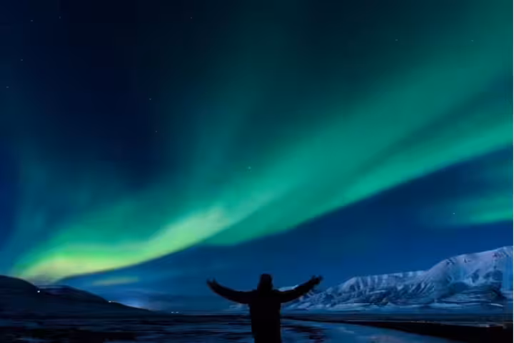 Person with arms raised watching vibrant green northern lights over snowy Iceland mountains at night