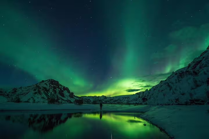 Person standing beneath vivid green aurora borealis over snowy Icelandic mountains reflected in still water