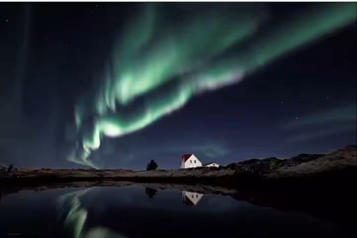 Aurora borealis dancing over a white house reflected in a calm lake in Iceland at night