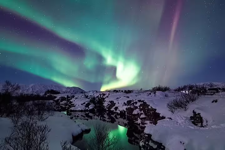 Vibrant green and purple northern lights dancing over snowy Iceland landscape with rocky terrain and still water reflection